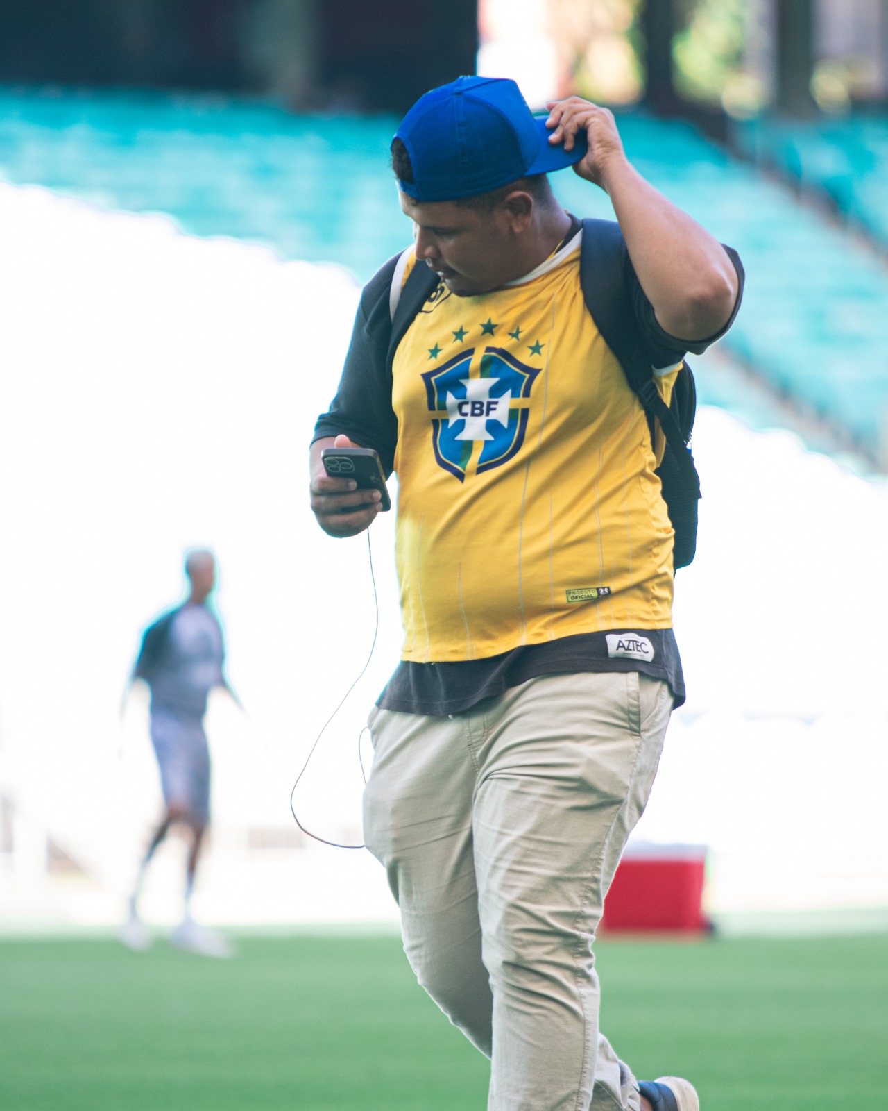 Samuel Moraes com colete CBF caminhando pelo gramado da arena com celular na mão